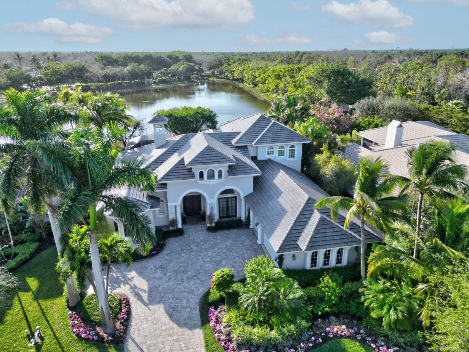 A luxury Naples estate featuring a newly installed dark grey concrete tile roof by Legrand's Roofing & Construction LLC, overlooking a lake with palm trees.