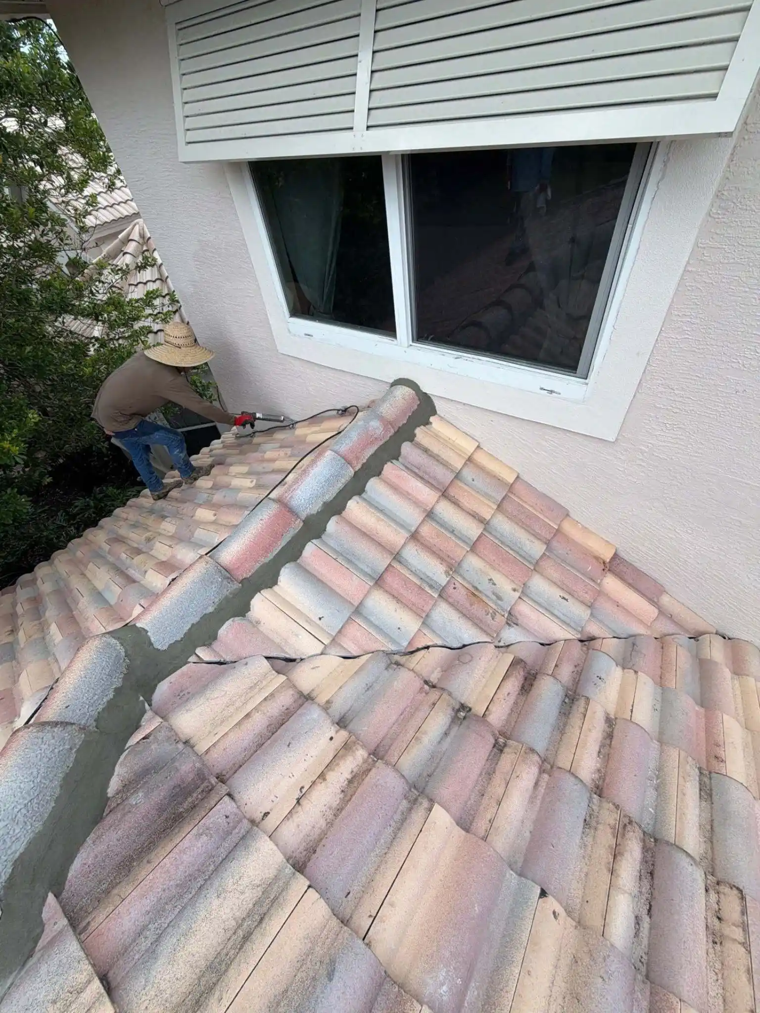 A professional roofer from Legrand's Roofing uses a trowel and mortar mix to reinforce a concrete tile hip ridge below a second-story window.