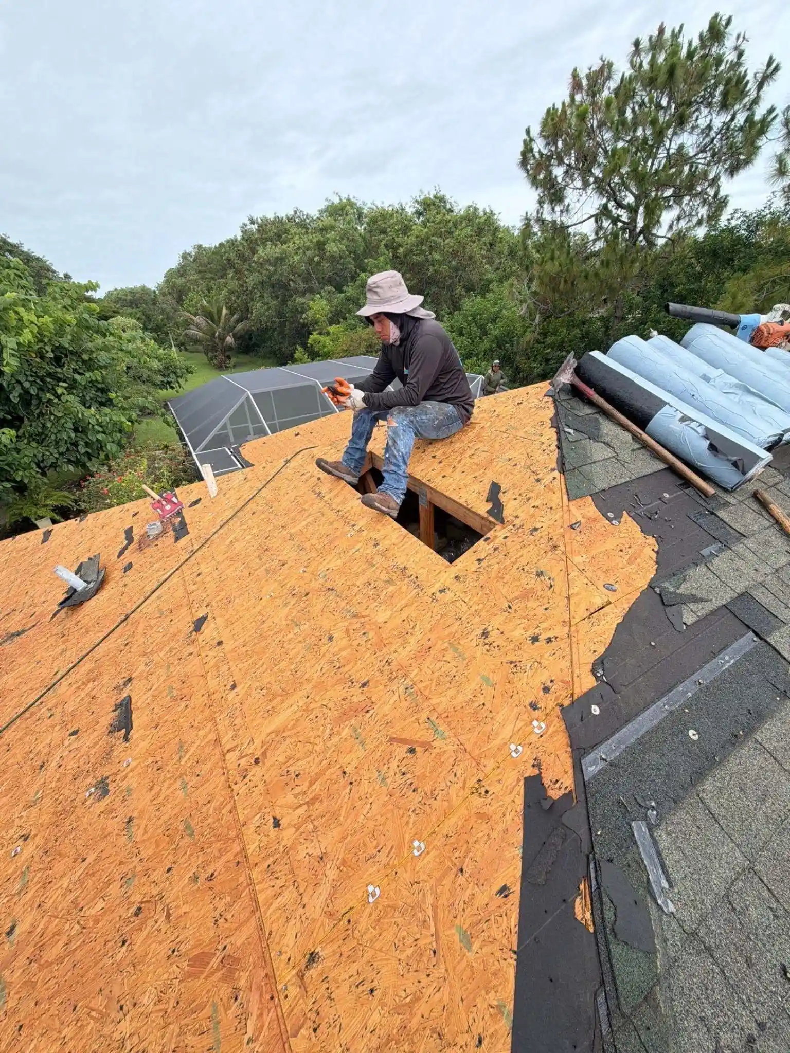 A Legrand's Roofing professional sits by an open framing cutout on a newly sheathed roof plane during a residential shingle reroof overhaul.