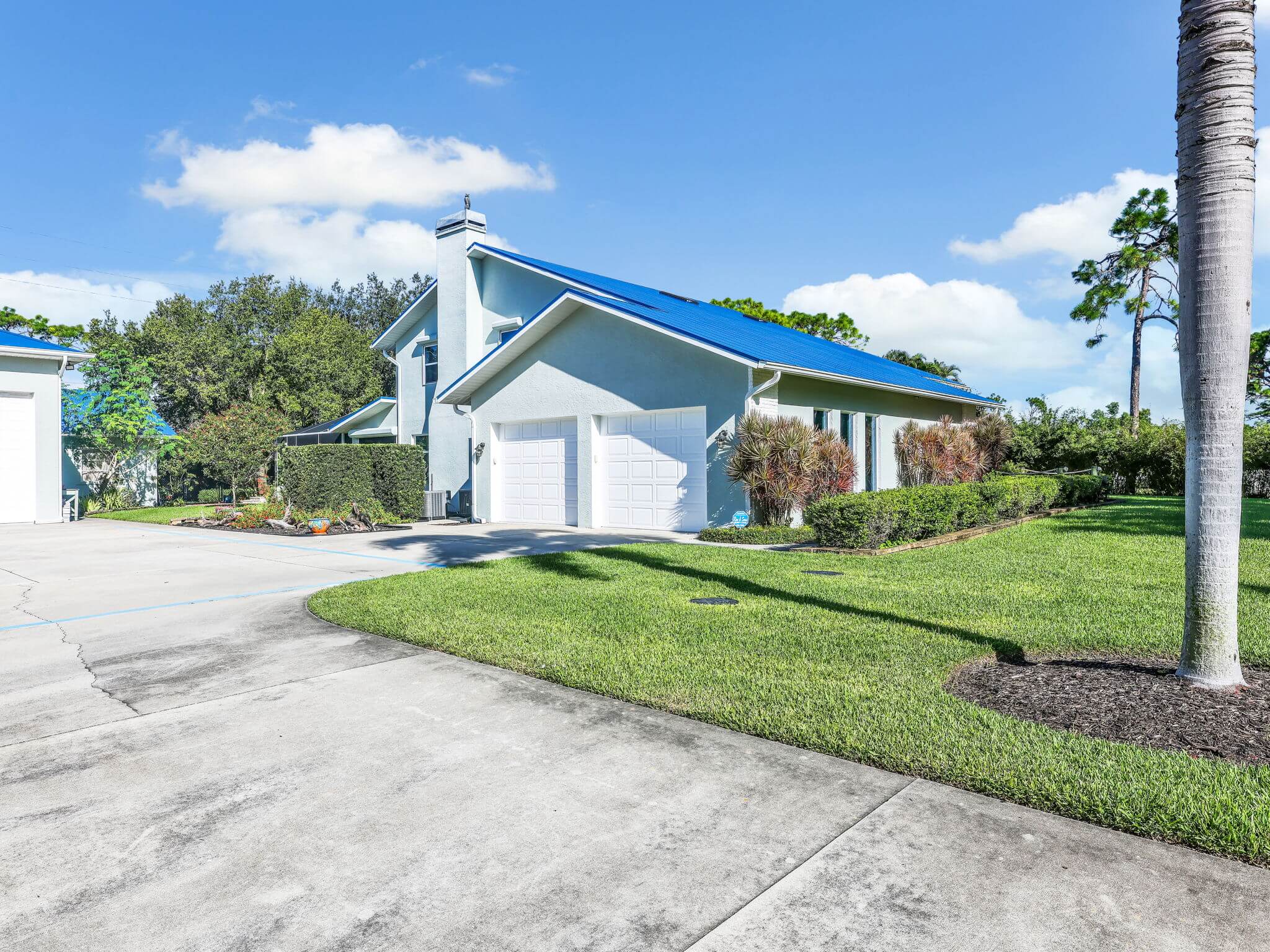 Driveway perspective photo showing the attached garage metal roof, chimney, and palm trees.
