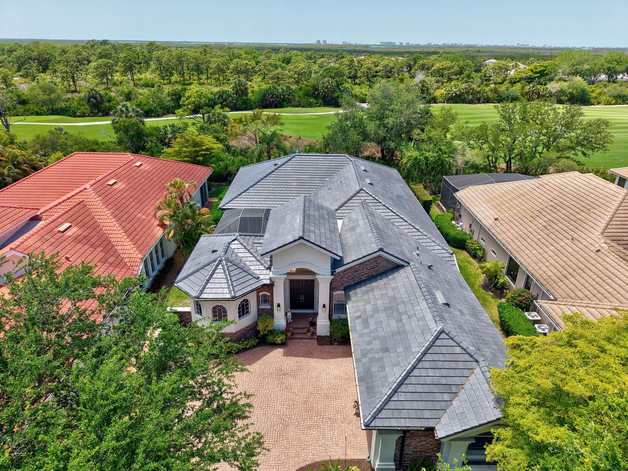 Elevated front view of the luxury estate home featuring dark flat tile roofing, paver driveway, and golf course backdrop.