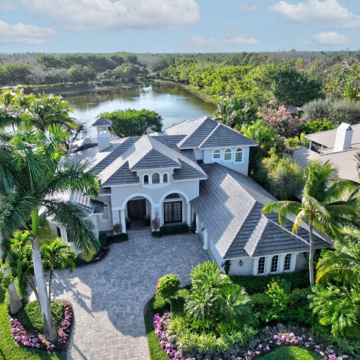 A luxury Naples estate featuring a newly installed dark grey concrete tile roof by Legrand's Roofing & Construction LLC, overlooking a lake with palm trees.
